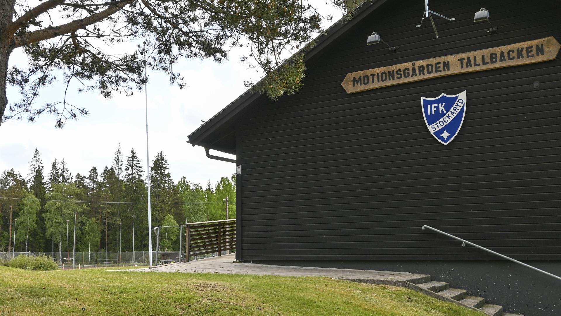 Brunsvart hus med texten Motionsgård tallbacken, träd och gräsmatta.